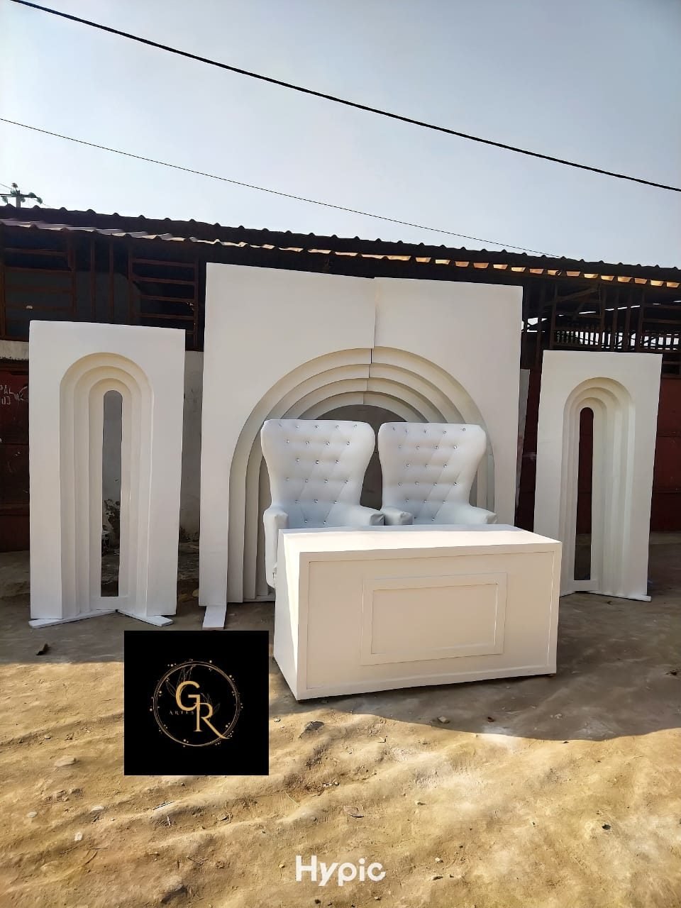 Cenário branco em madeira com arco decorativo ao fundo, duas cadeiras estofadas tipo trono e uma mesa branca à frente.