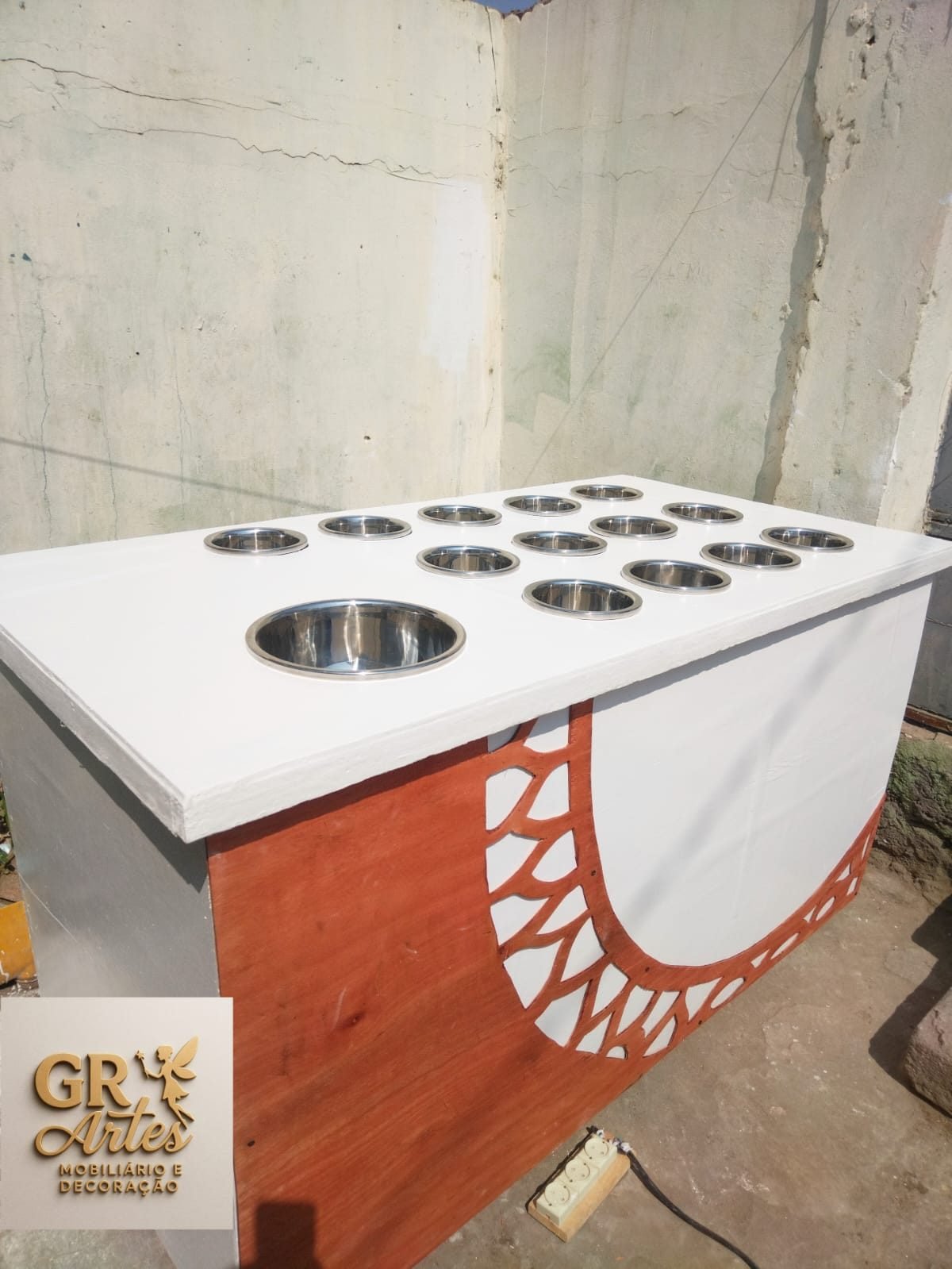 Mesa buffet branca com catorze cubas redondas em aço inoxidável embutidas no tampo e um painel lateral vazado em laranja e branco.