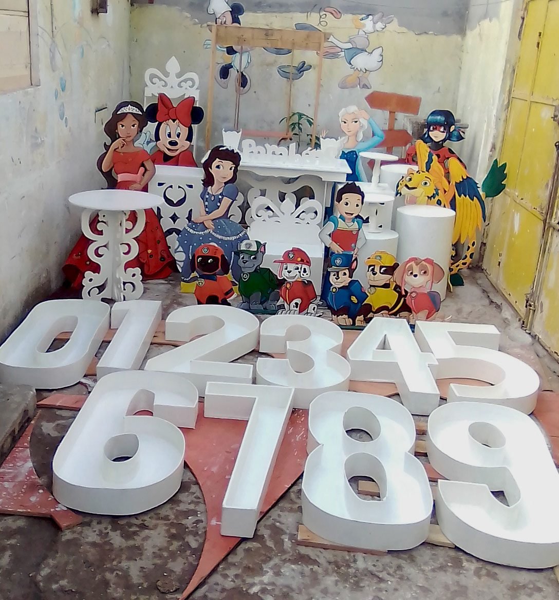 Conjunto de números grandes de zero a nove e figuras de personagens infantis recortados em madeira, dispostos no chão.